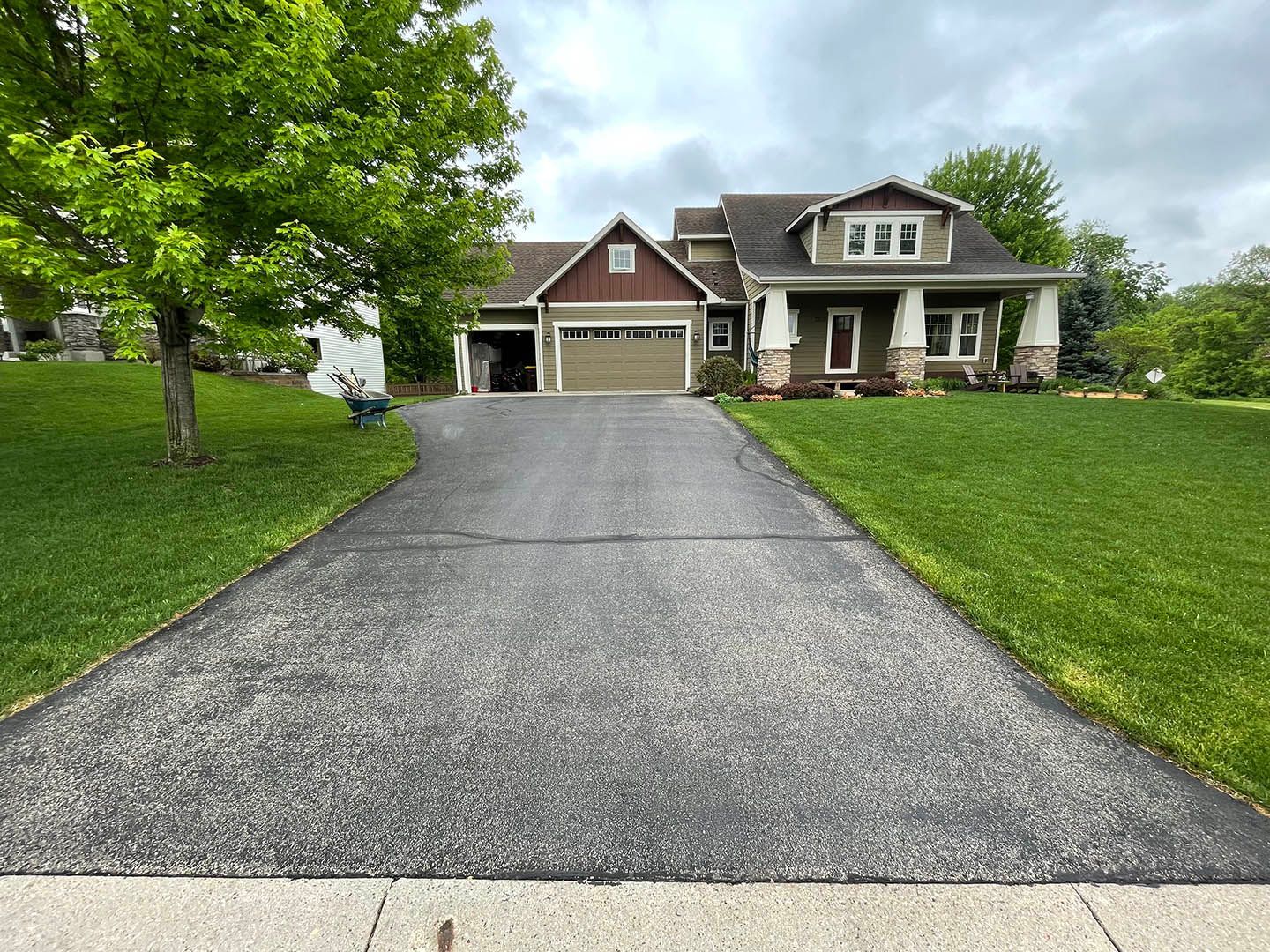 A two-story suburban home with a dark roof, tan siding, and stone accents, viewed from a paved driveway under cloudy skies.