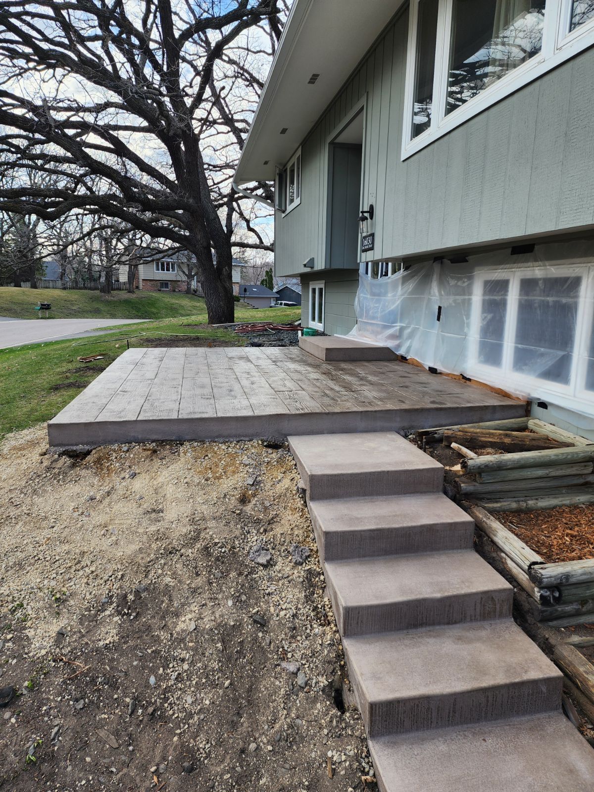 A new gray concrete patio and stepped entrance leading to the front door of a house with a large tree in the yard.