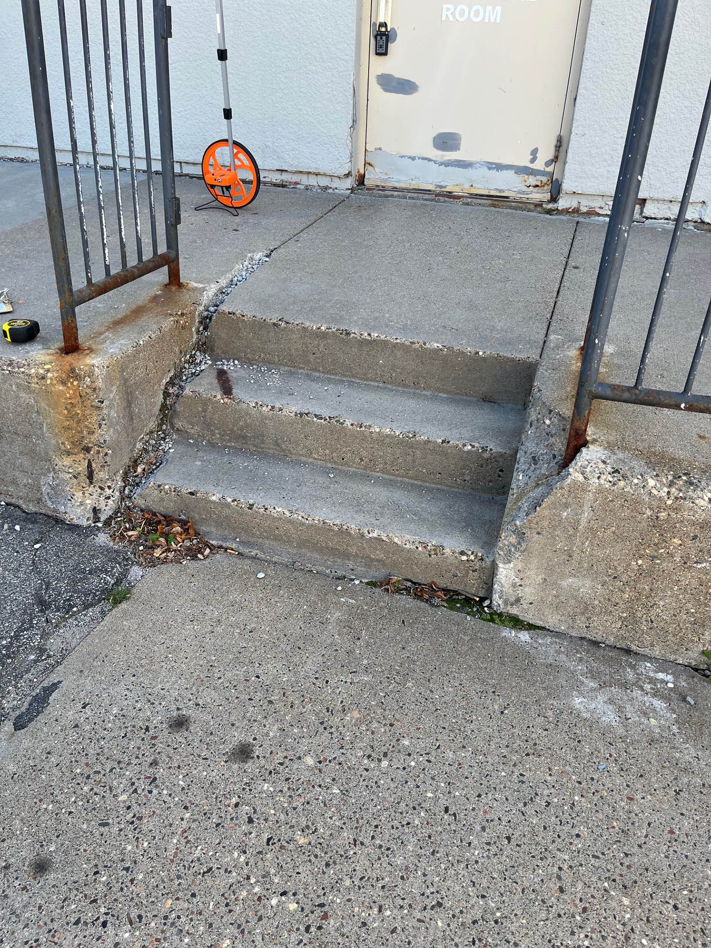A set of two concrete steps leading to a building door, flanked by metal railings with visible rust and crumbling concrete.
