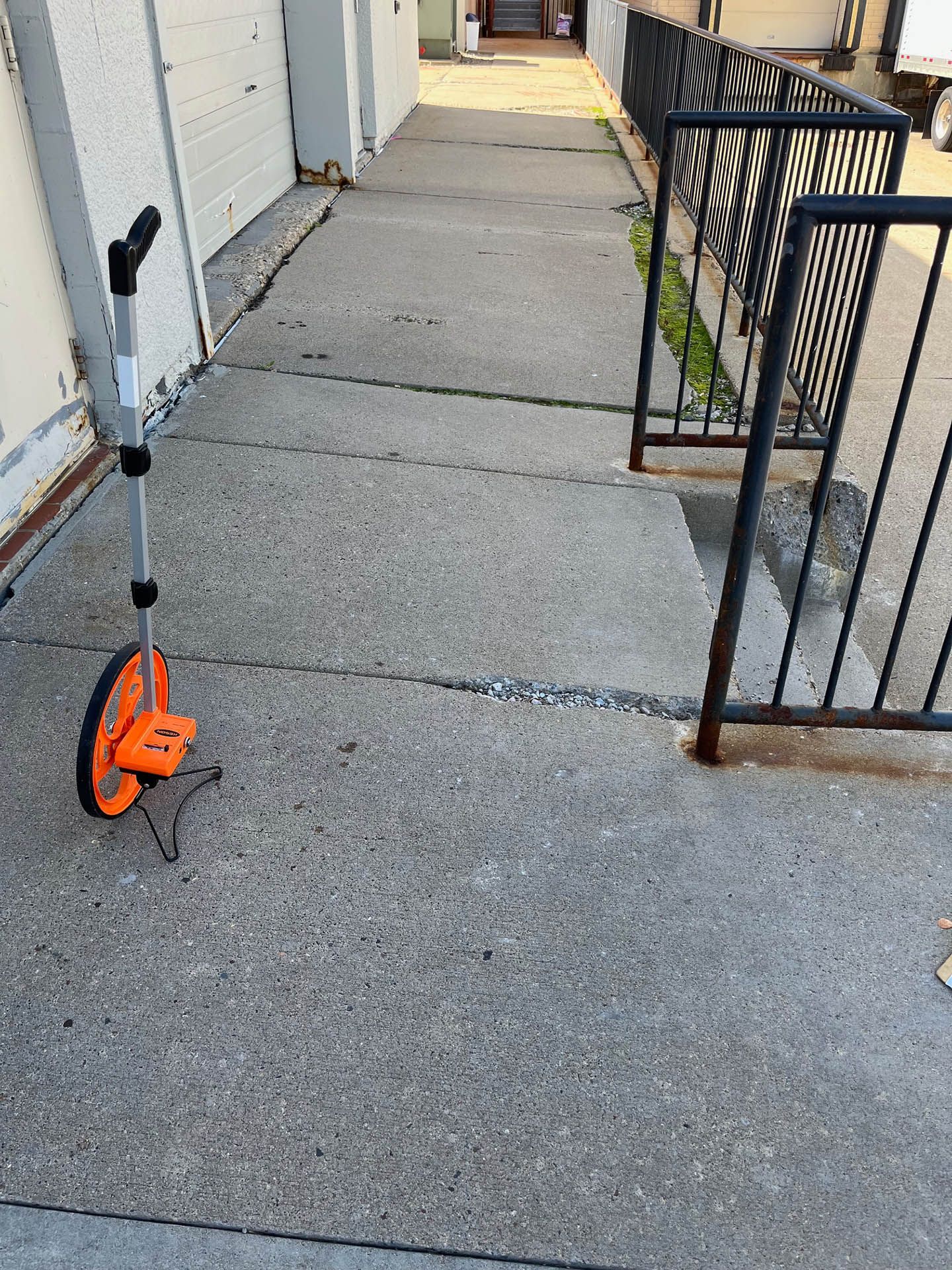 An orange distance measuring wheel stands on a concrete sidewalk beside a black metal railing.