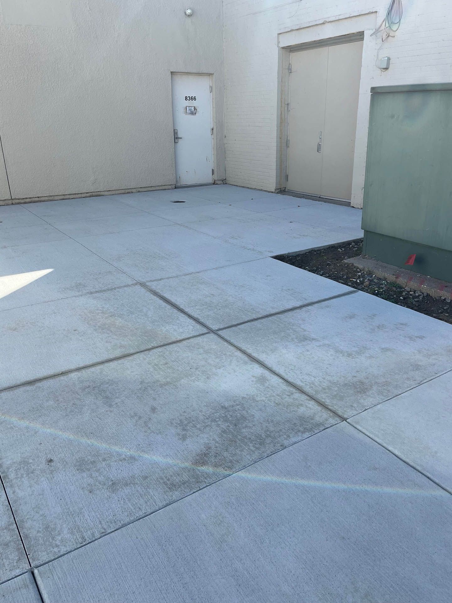A paved exterior area featuring a light-colored building, a white single door, and a large double door next to a green box.
