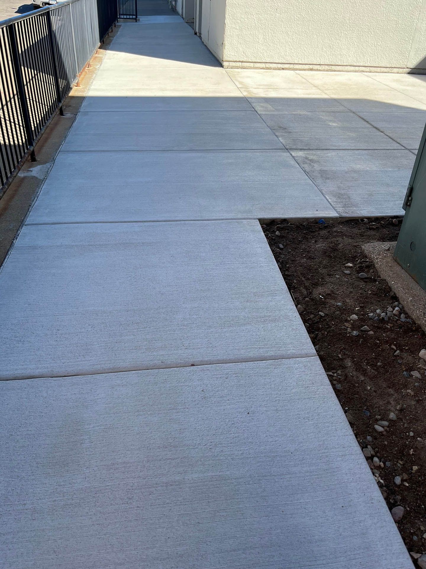 A paved concrete walkway with expansion joints, bordered by a black metal fence on the left and a dirt patch on the right.