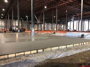 A large industrial warehouse under construction featuring a newly poured concrete floor and steel support columns.