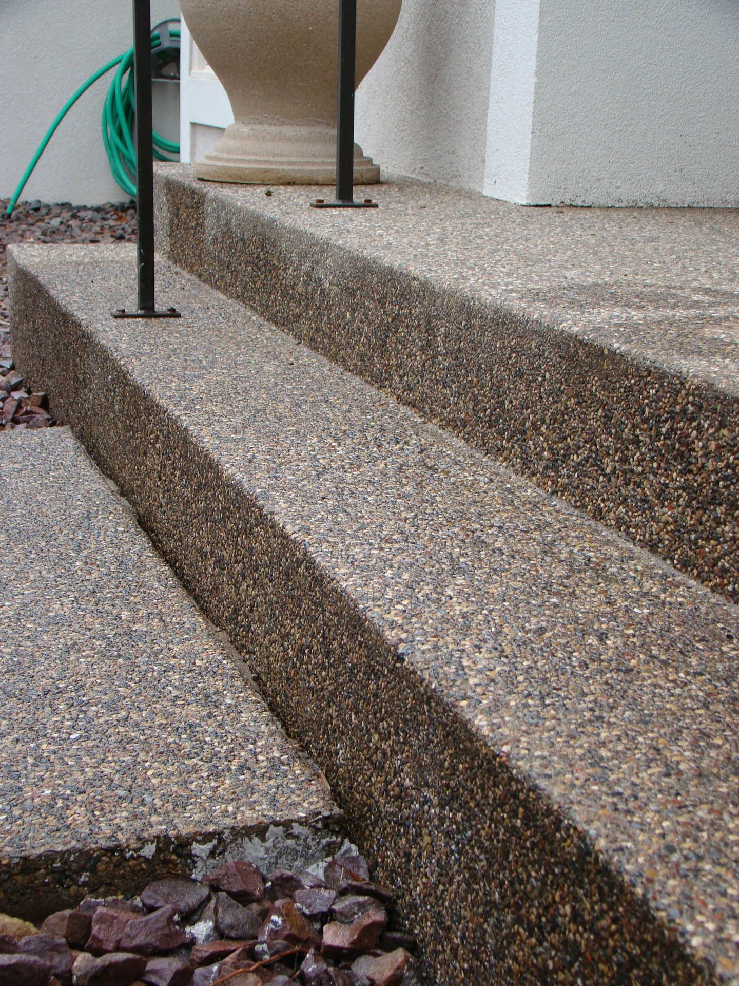 A high-angle view of two concrete steps with an exposed aggregate finish, adjacent to a white wall and a stone planter.