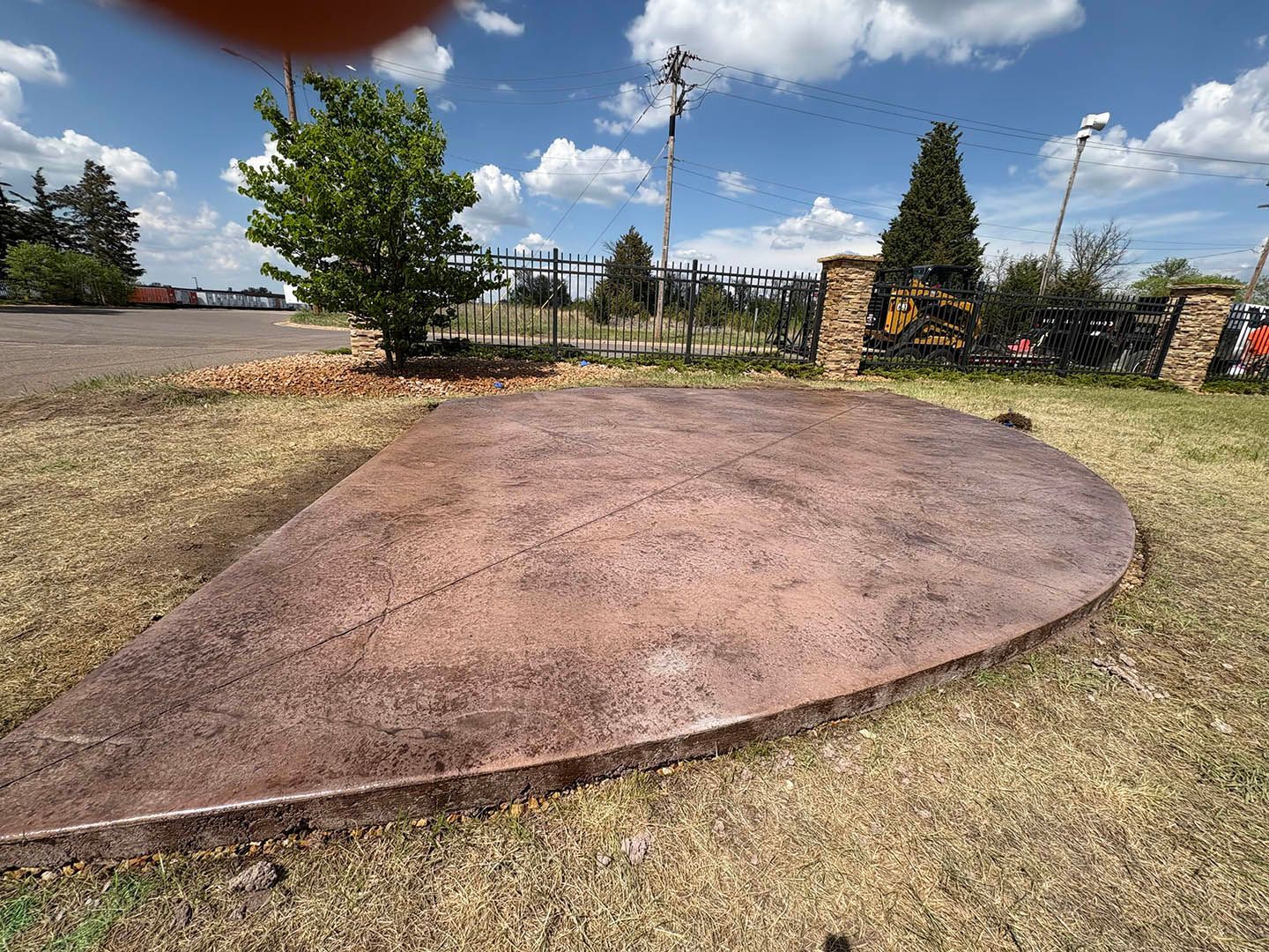 A semi-circular, stamped concrete patio with a mottled reddish-brown finish set in a grassy yard under a blue sky.