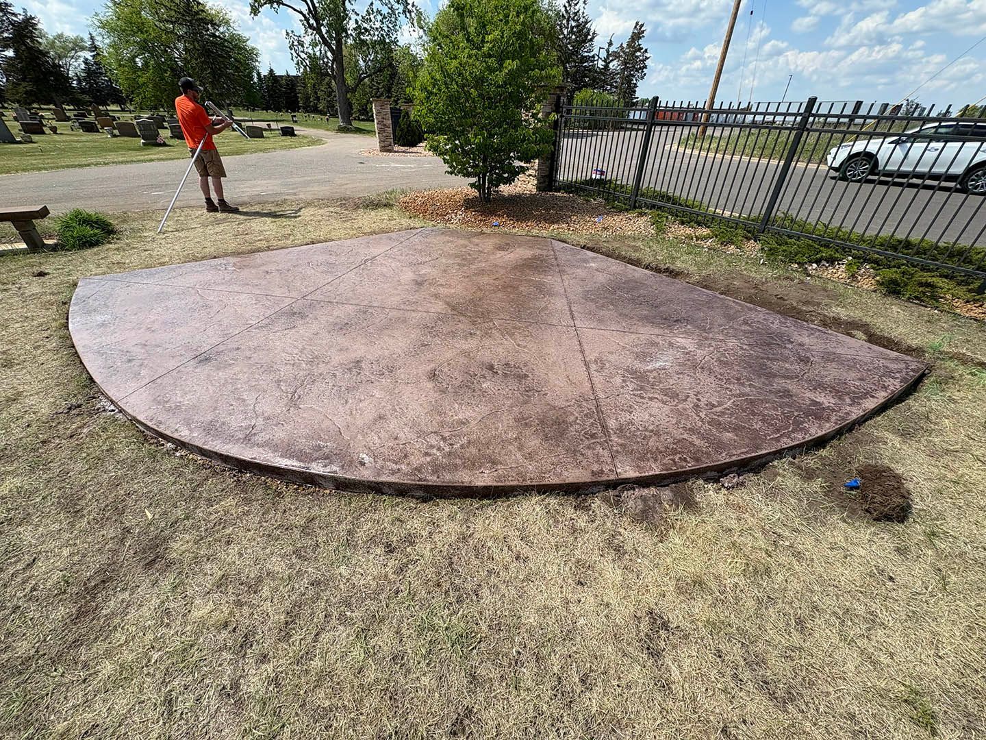 A person stands near a circular, stained-concrete patio set in a grassy area with a cemetery in the background.