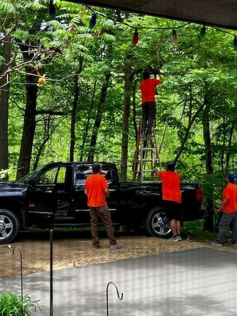Four people in bright orange shirts work on setting up outdoor string lights in a wooded area near a black pickup truck.