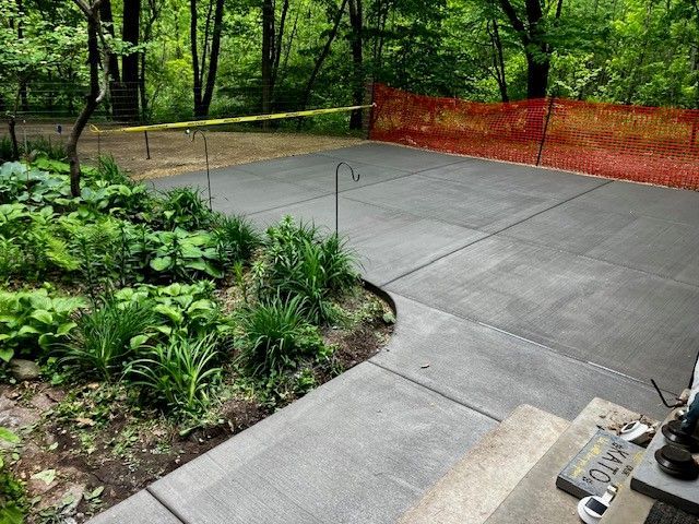 A newly poured concrete patio edged with garden plants, bordered by yellow tape and an orange construction fence.