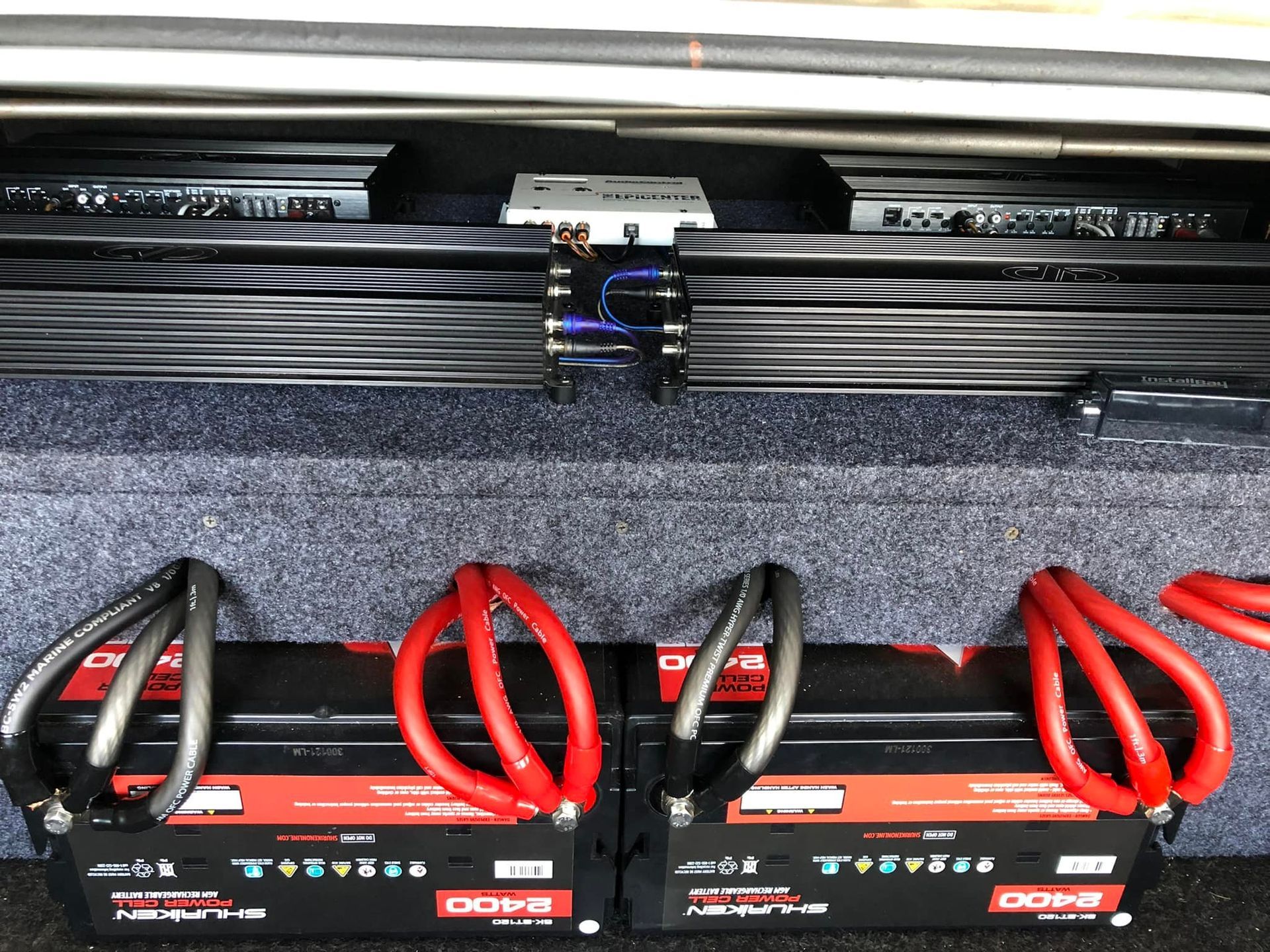 Two amplifiers, two car batteries, black and red cables, carpeted enclosure.