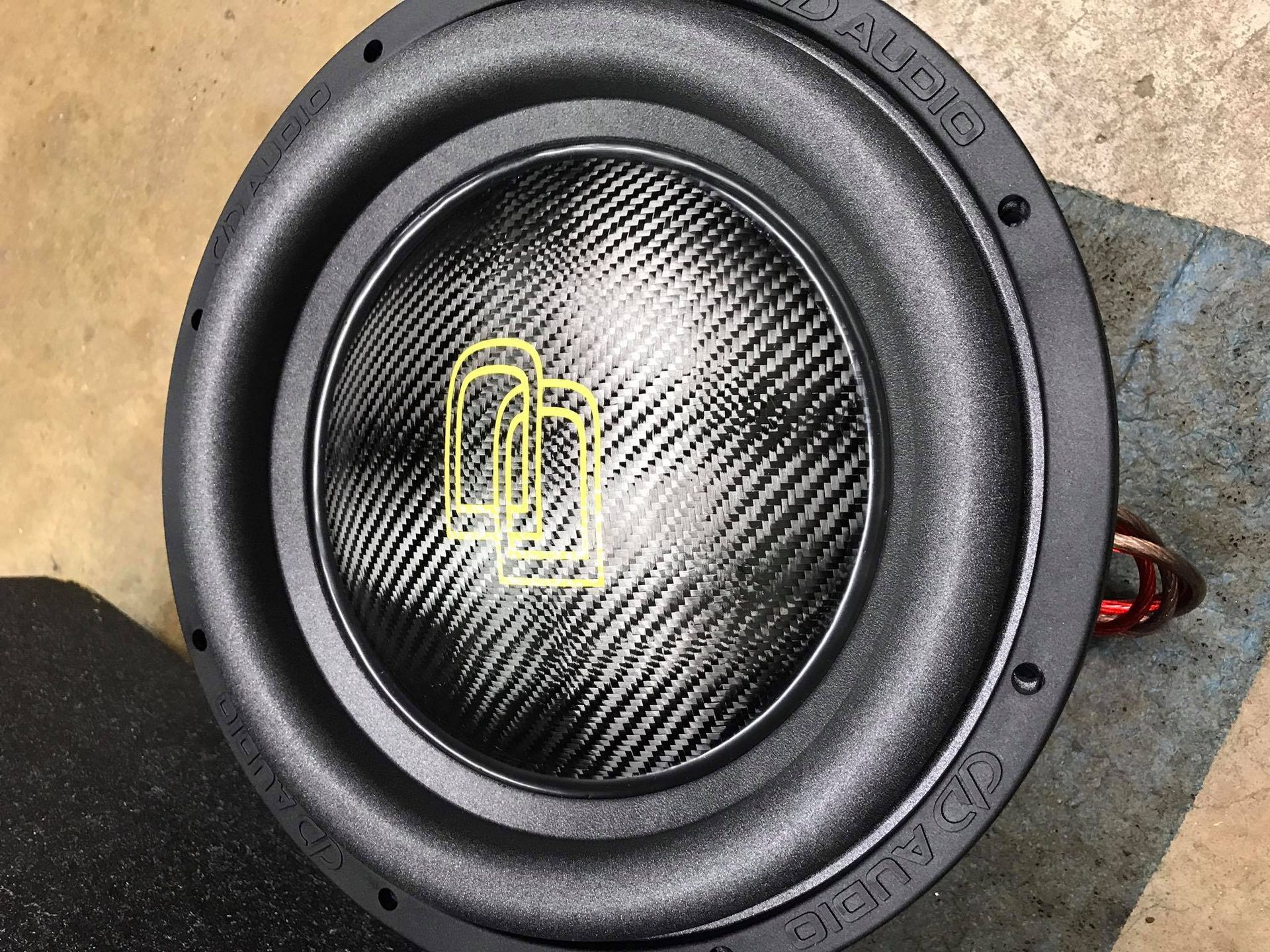 Black subwoofer with carbon fiber cone and yellow logo, on a gray surface.