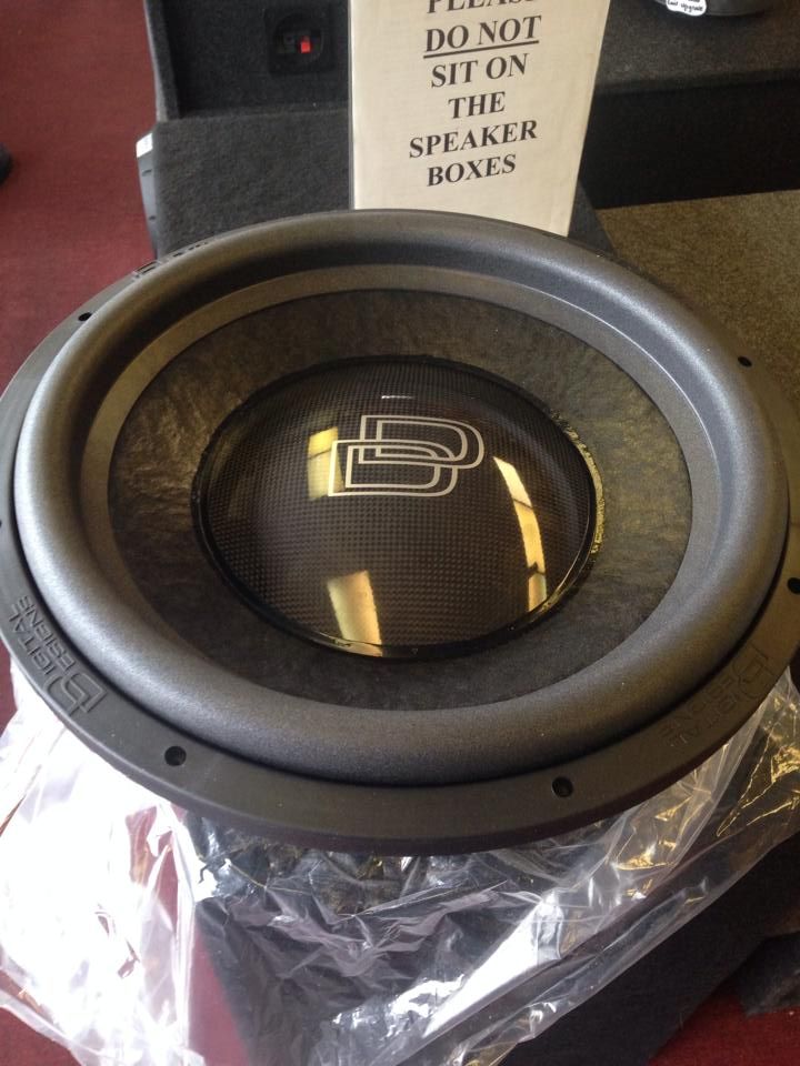 Black subwoofer with a mesh screen and logo, sitting on plastic wrap, in front of a sign that says