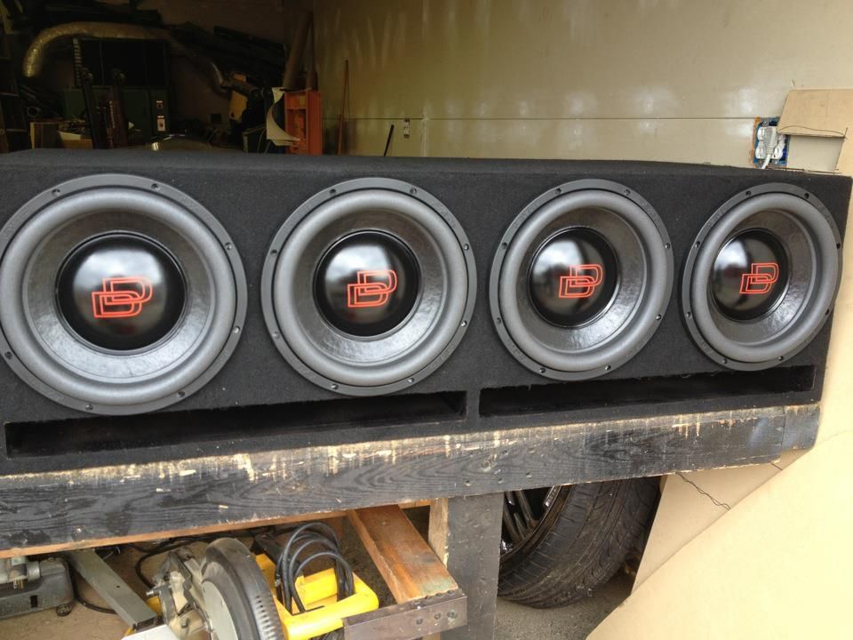 Four black car subwoofers in a custom enclosure, mounted in the bed of a vehicle.