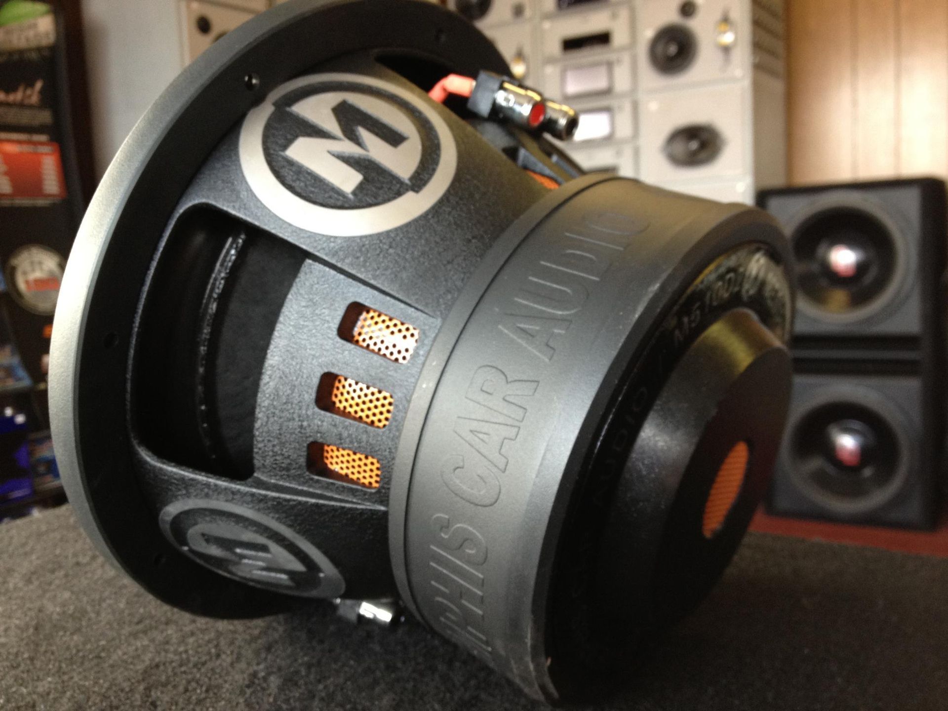 Close-up of a black MISCAR AUDIO subwoofer with orange accents, in a car audio shop setting.