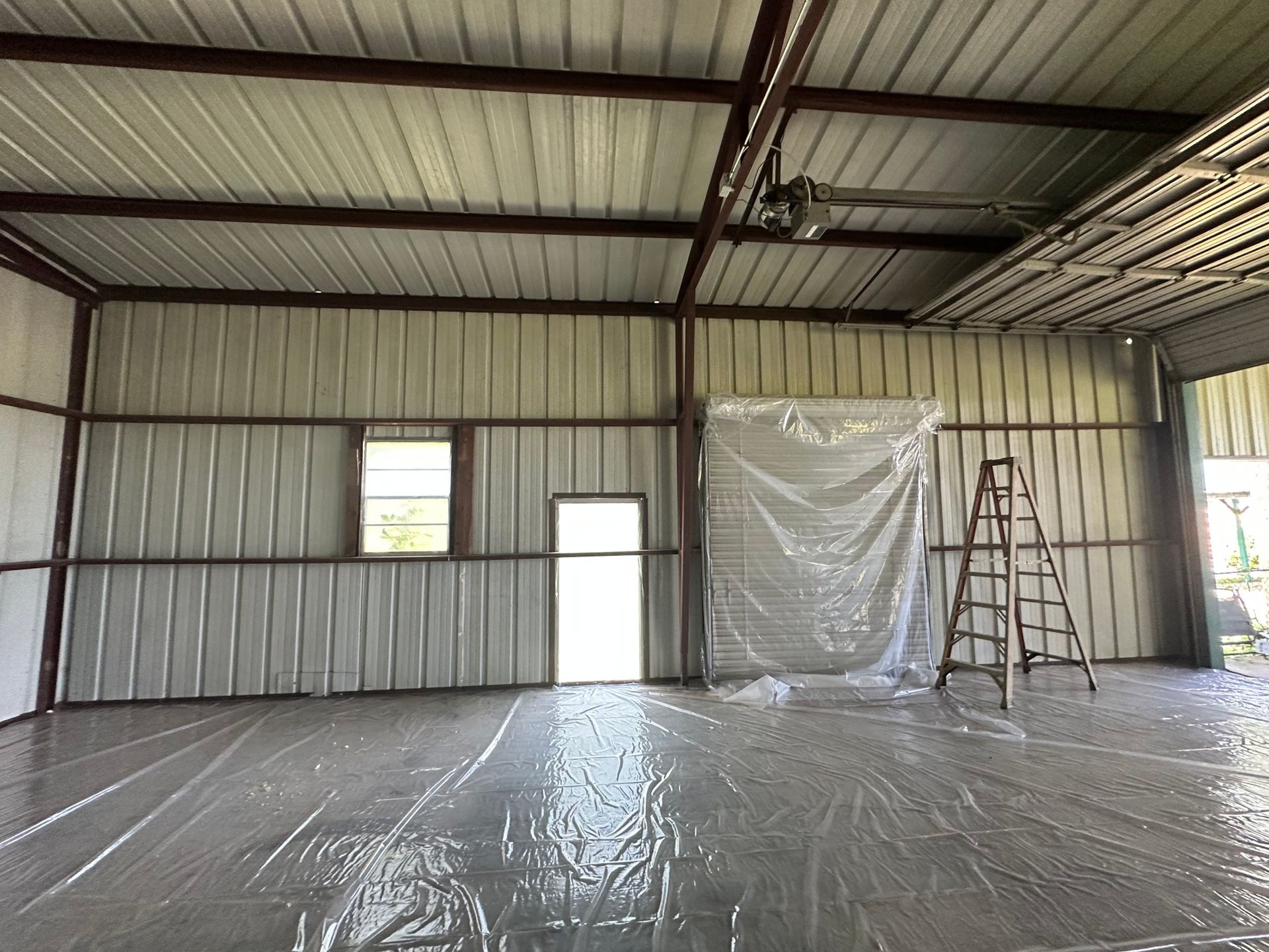 Inside of a metal-walled building with a plastic sheet covering the floor. Includes a door, windows, and ladder.