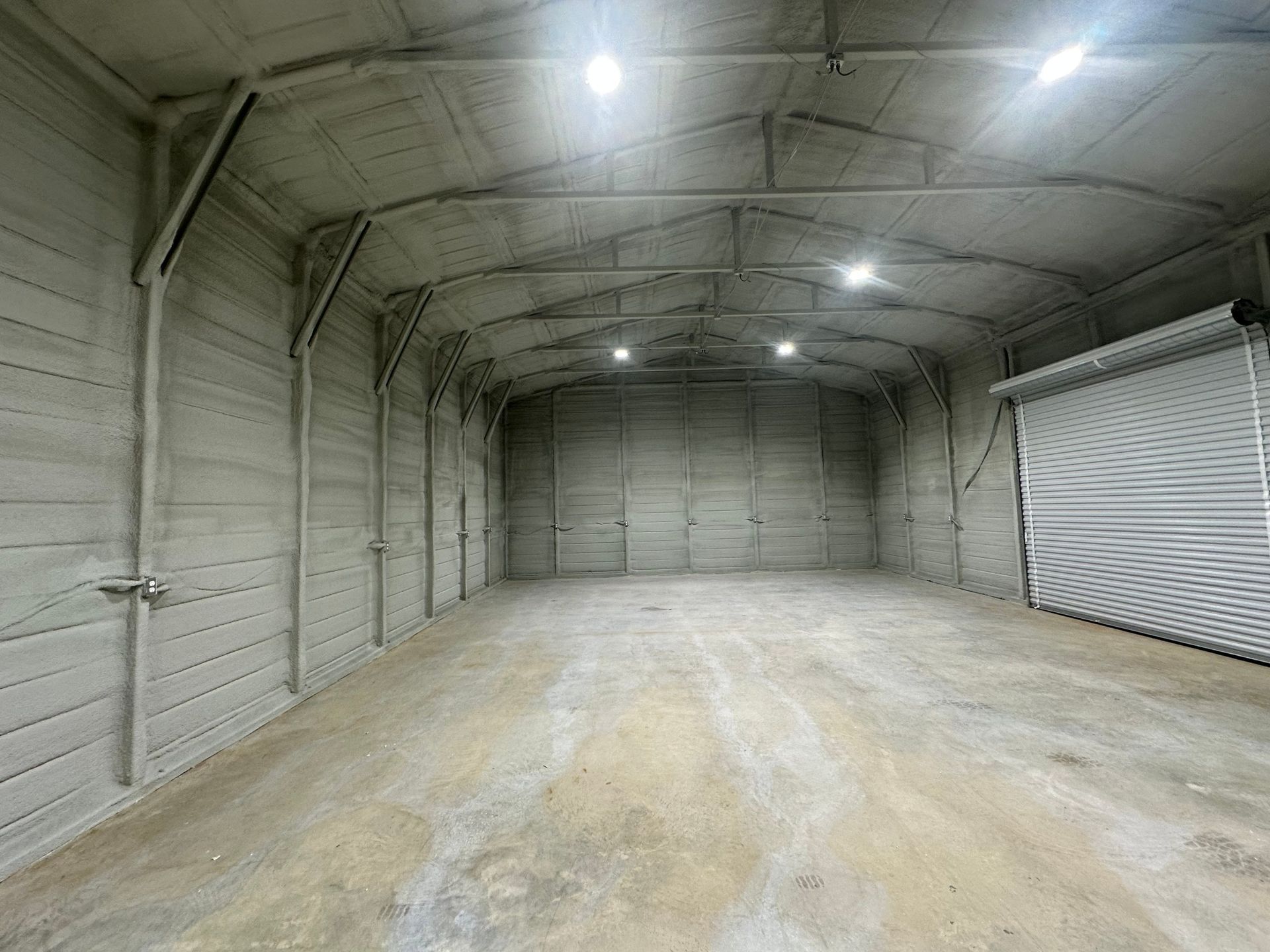 Interior of an empty concrete warehouse with a roll-up door, lit by overhead lights.