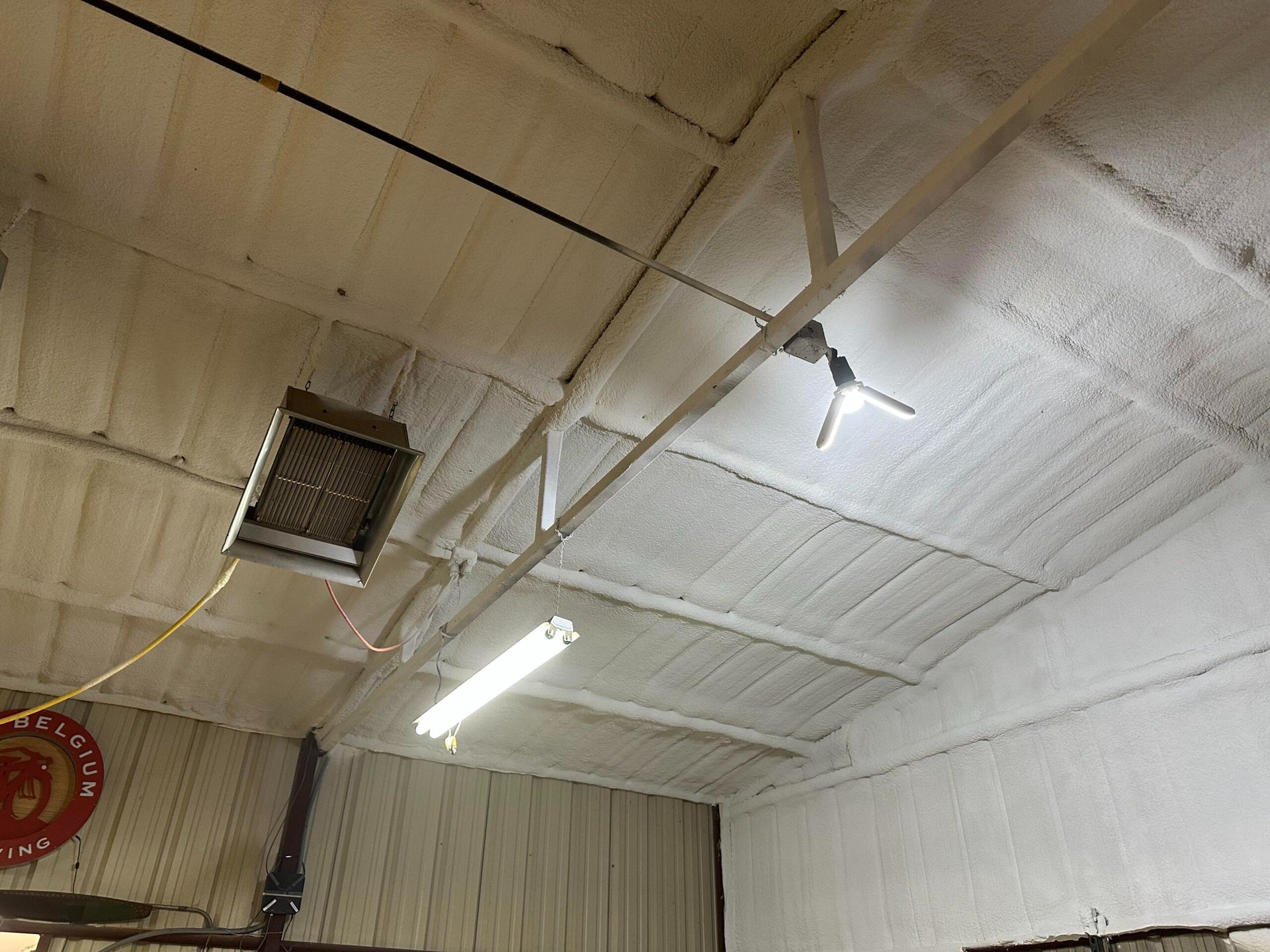 Foam insulation on a garage ceiling with beams, a heater, light fixtures, and a ceiling fan.