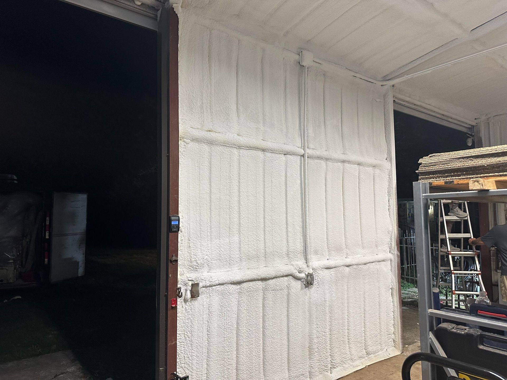 White insulation sprayed on a metal wall inside a building. Open door on the left; ladder on the right.