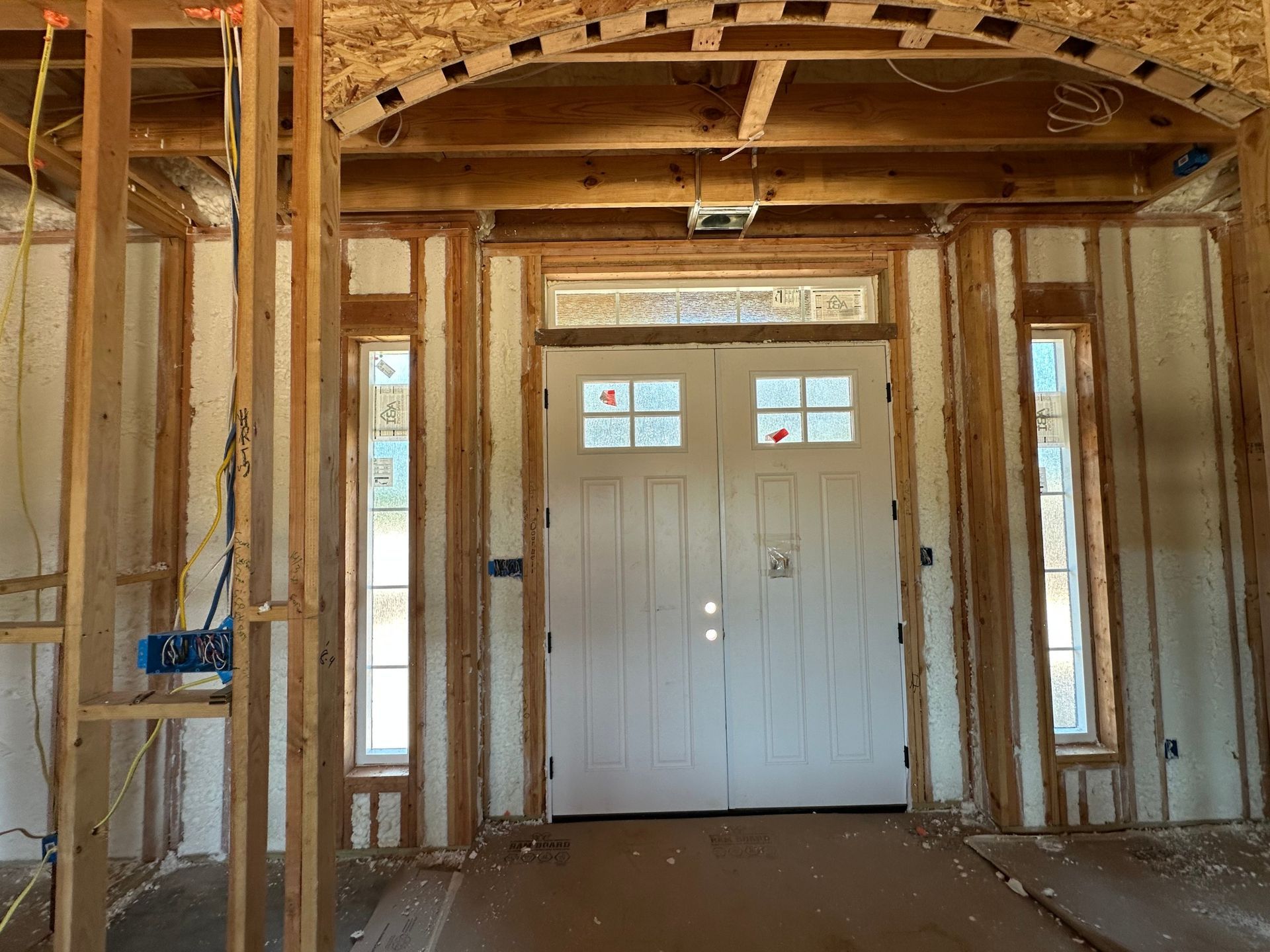 Interior view of a building under construction, featuring a white double door flanked by windows and wood framing.