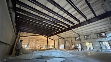 Interior of a building under construction, steel beams, spray foam insulation, workers, and a large roll of material.