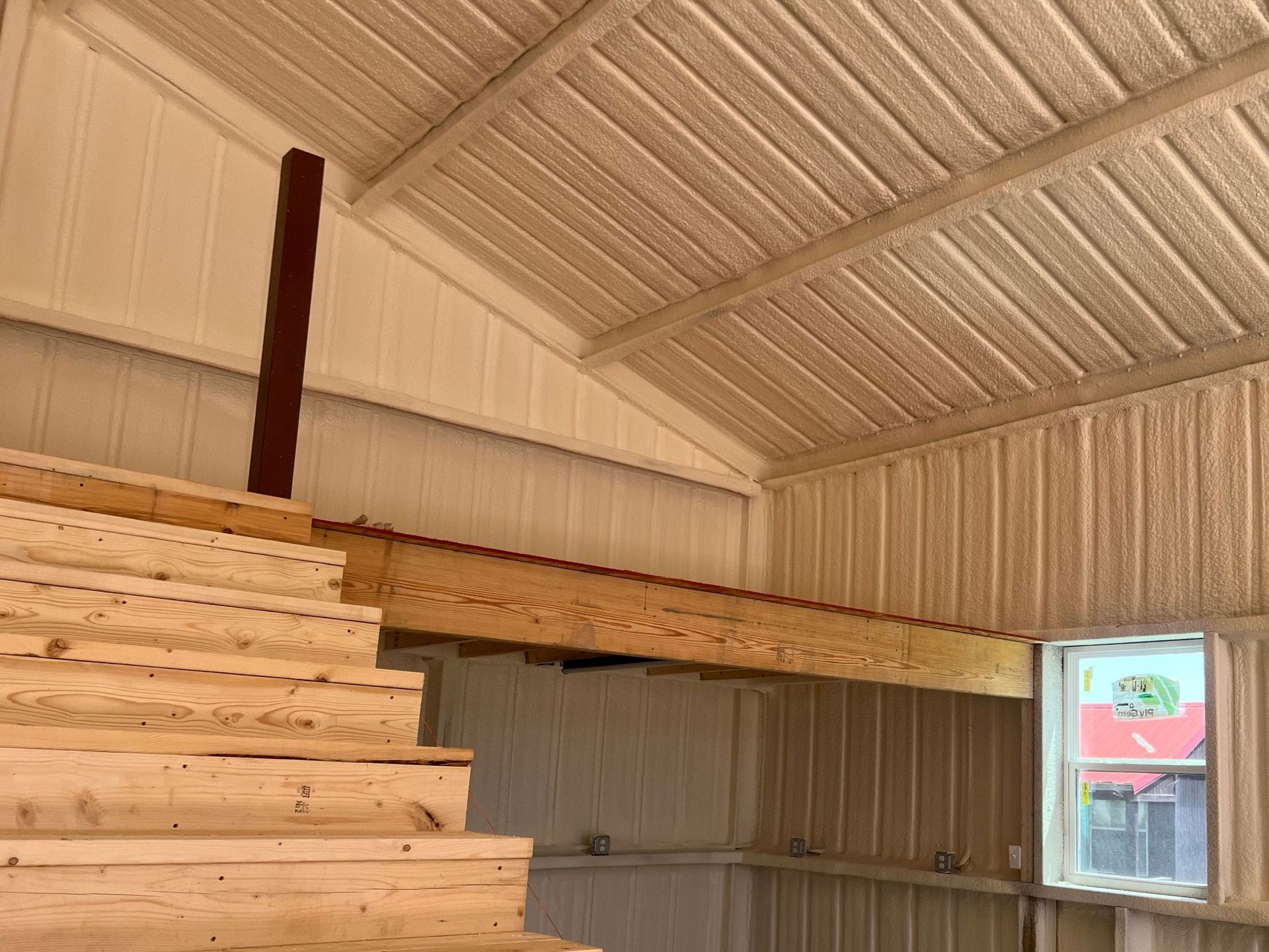 Interior view of a loft with a wooden staircase and a window. Light-colored walls and ceiling are visible.