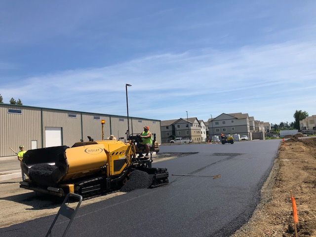 A yellow tractor is paving a road in front of a building.