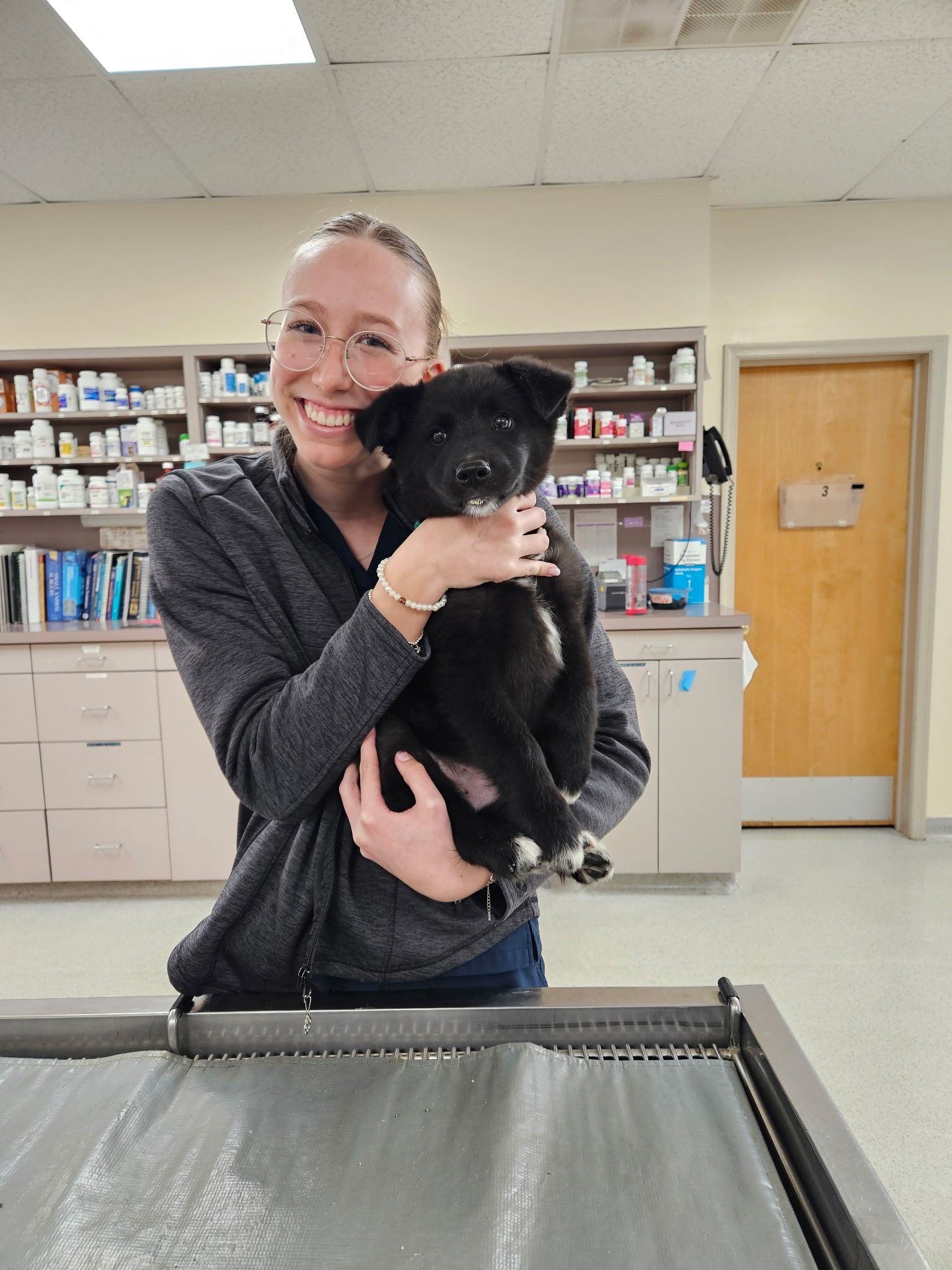 Woman smiling, holding a black puppy with white paws, in a veterinary clinic exam room.