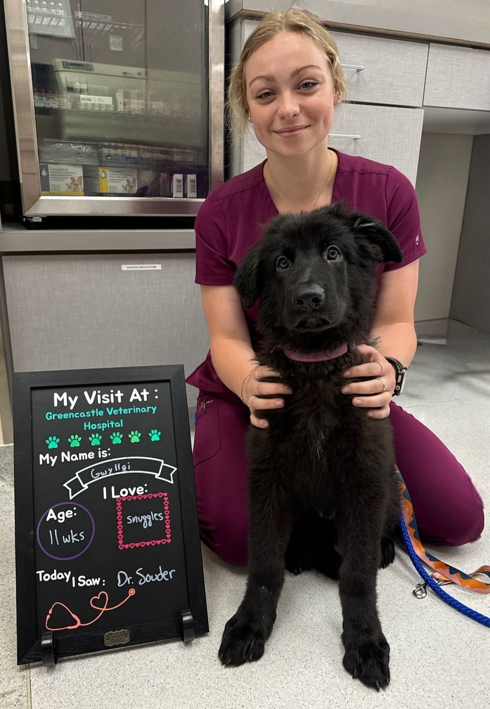 Veterinary tech and black puppy pose with visit sign at a clinic.