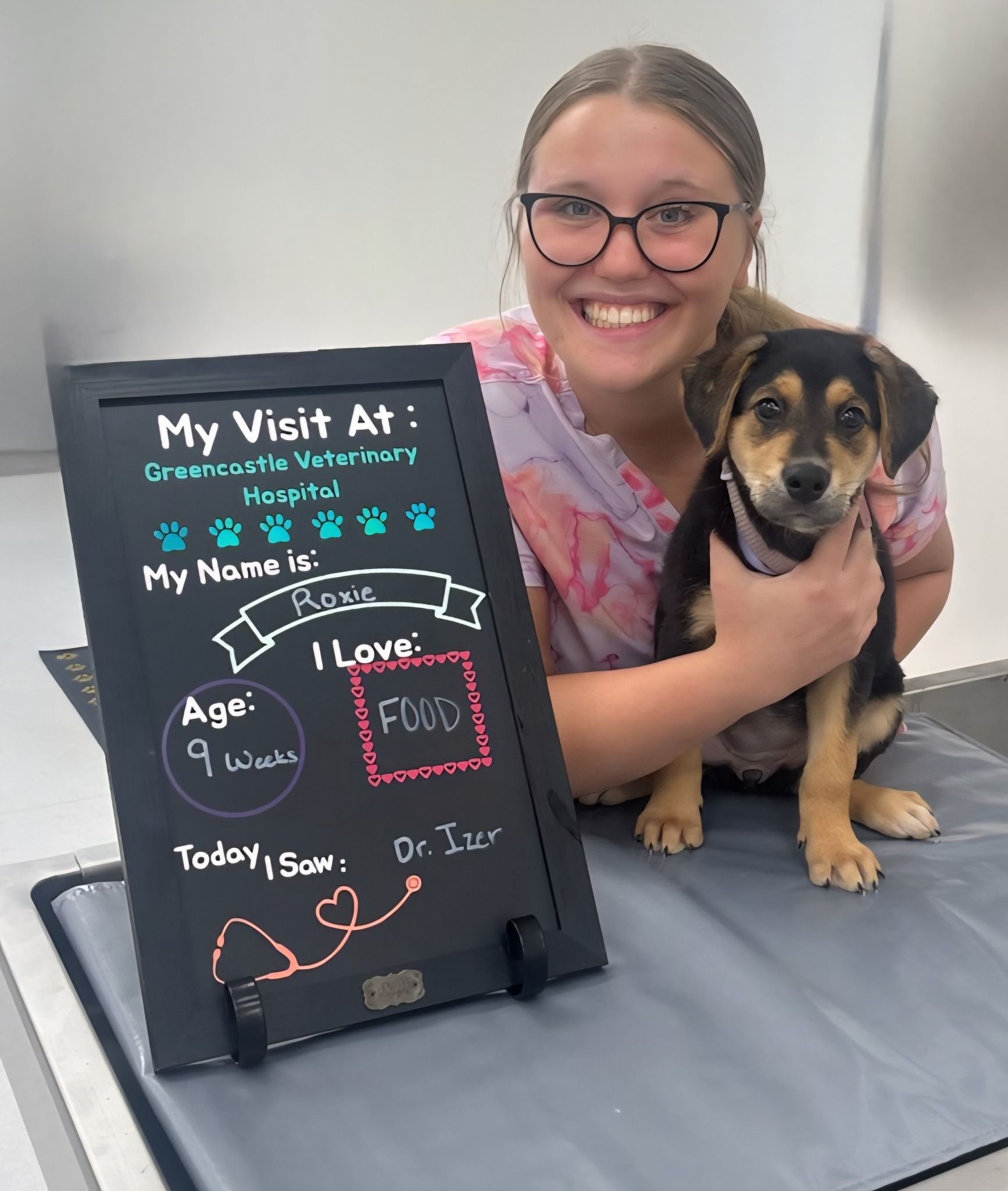 Woman with puppy poses beside a chalkboard. Chalkboard says 