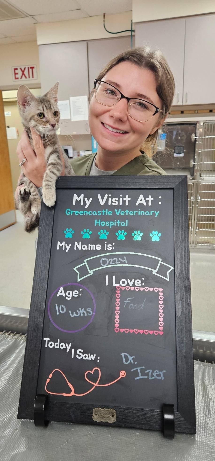 A smiling woman holds a kitten at a veterinary office. A chalkboard indicates the cat’s visit.