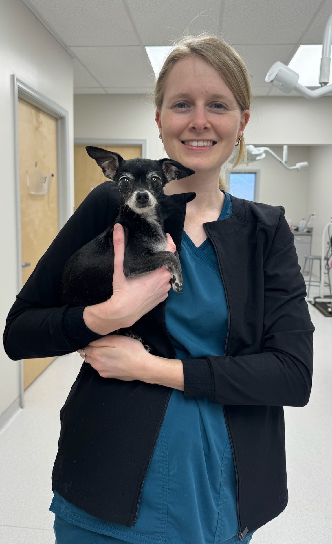Woman in teal top and black jacket holds a small black dog in a veterinary clinic.