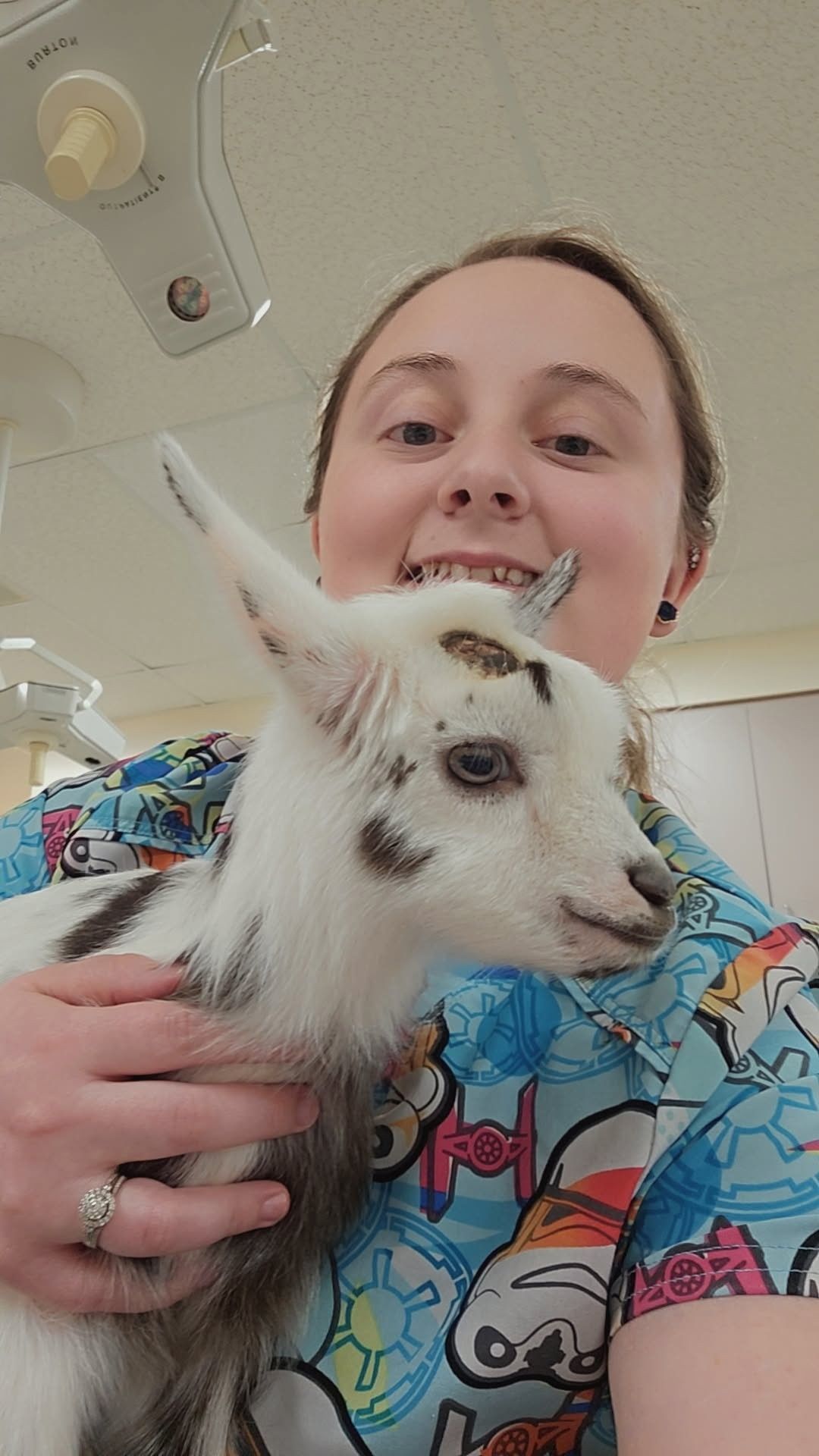 Woman holding a small goat, both smiling. The setting appears to be a veterinary clinic.