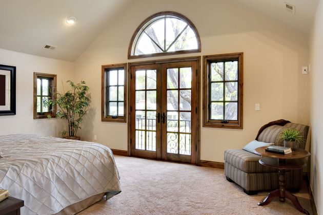 Bright bedroom with French doors, arched window, beige walls, and a wicker chair by the window.