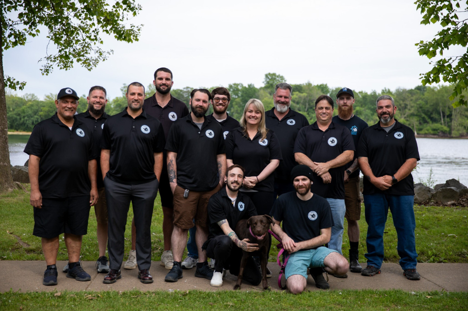 Group of people in black shirts posing outdoors by a river, with two dogs in front.