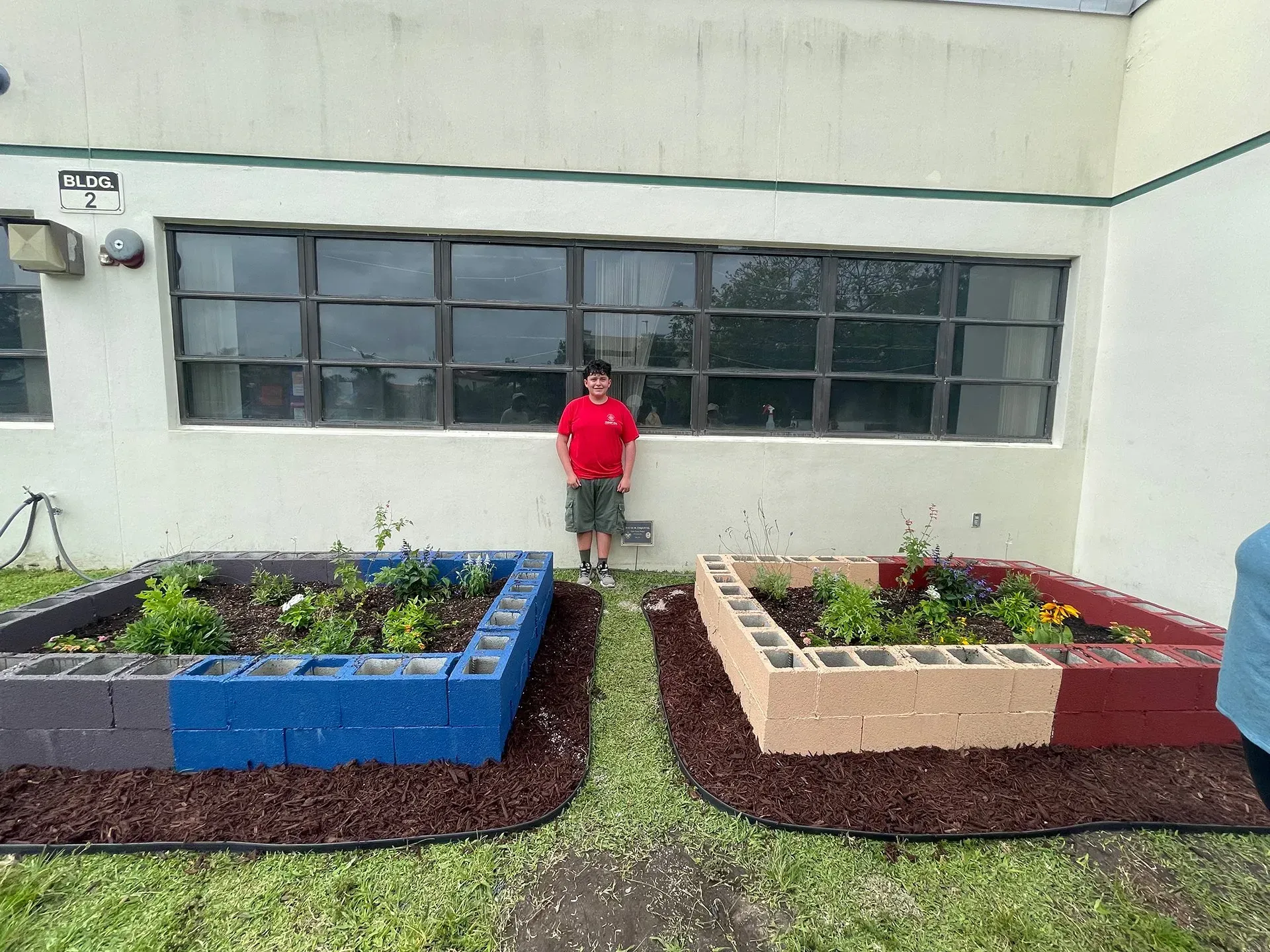 A boy is standing in front of a building next to a garden.