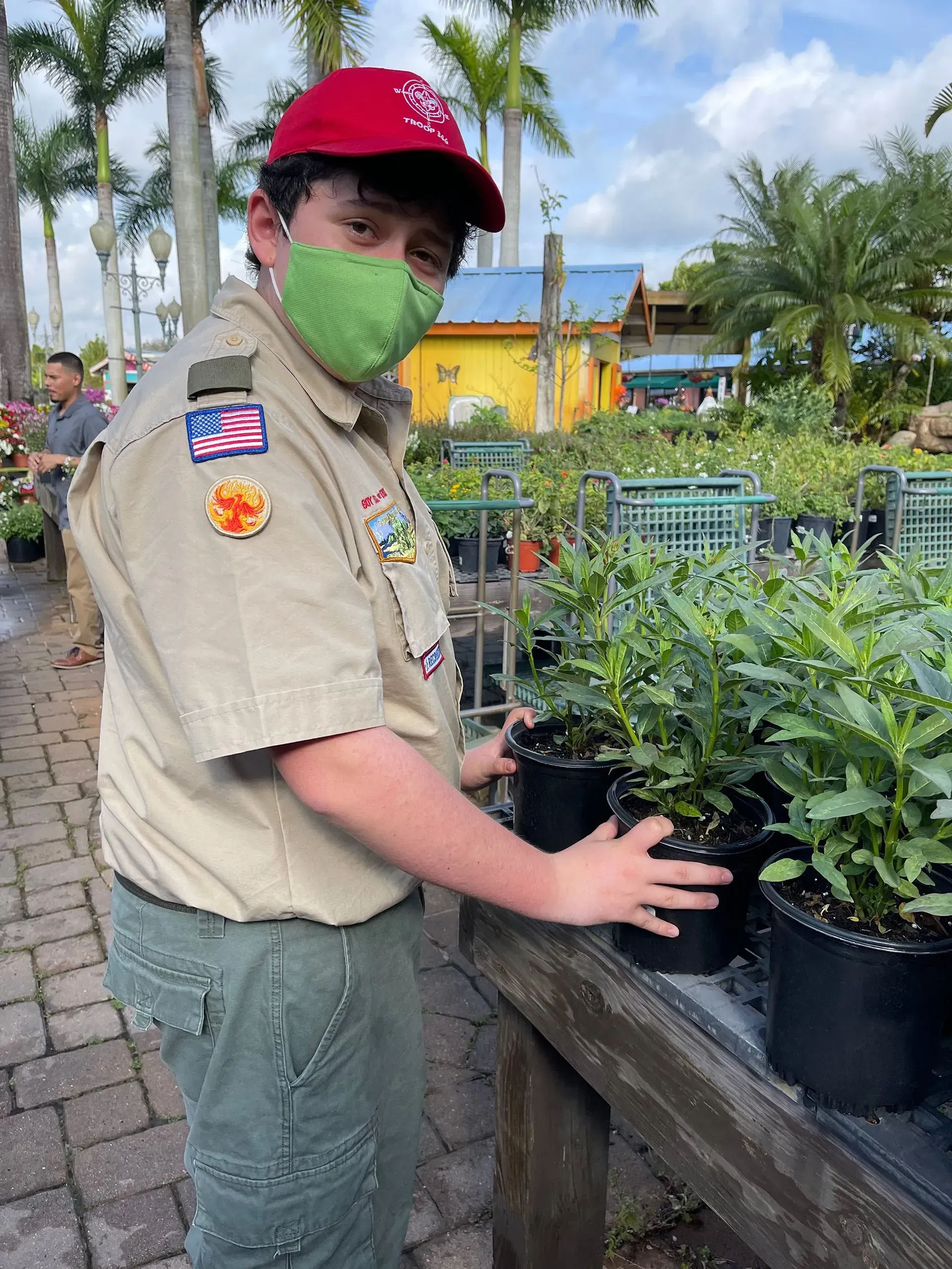 A boy scout wearing a green mask is holding a potted plant.