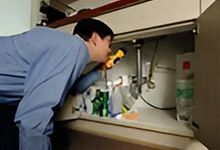 Person in blue shirt looking under a sink with a flashlight.