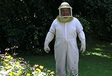 Person in white beekeeping suit stands in a grassy area with flowers and foliage.