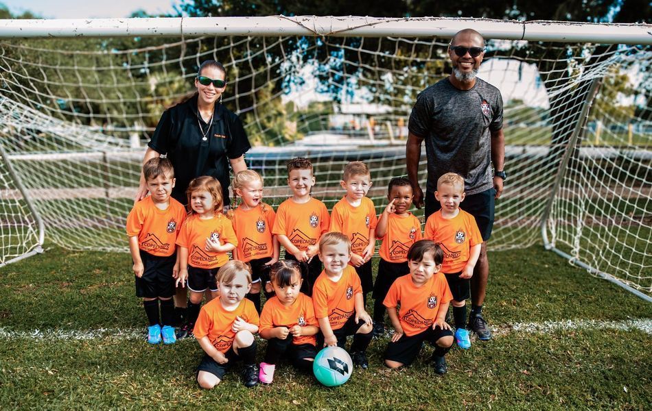 Youth soccer team in orange jerseys poses with coaches in front of a goal.