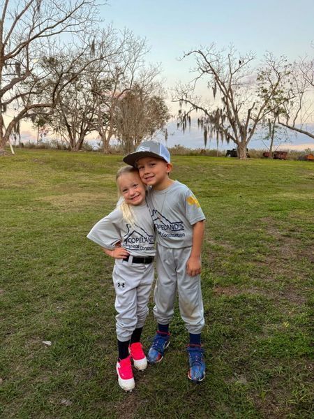 Two young children in baseball uniforms, posing for a photo outdoors.