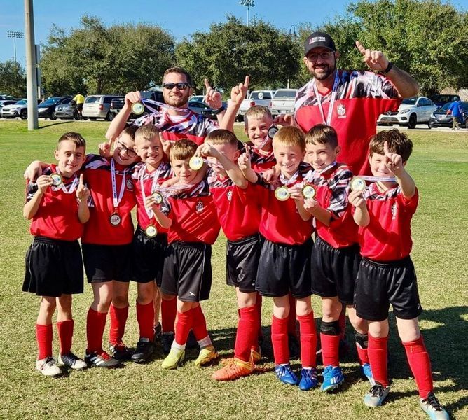 Youth soccer team in red and black uniforms celebrating a victory, holding medals, on a grassy field.