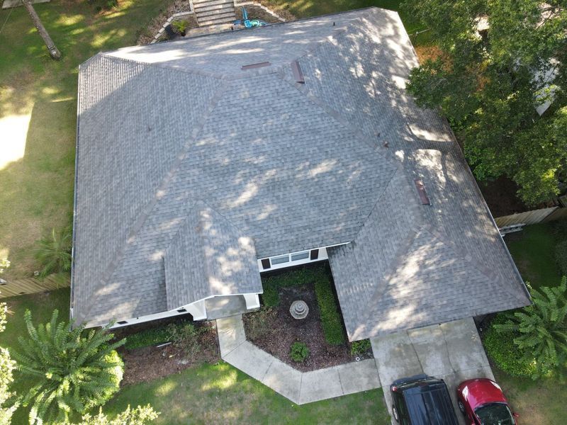 Aerial view of a house with a gray shingle roof, driveway, and green landscaping.