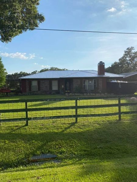 A brick ranch-style house with a metal roof behind a green fence and lawn, trees, and a clear sky.