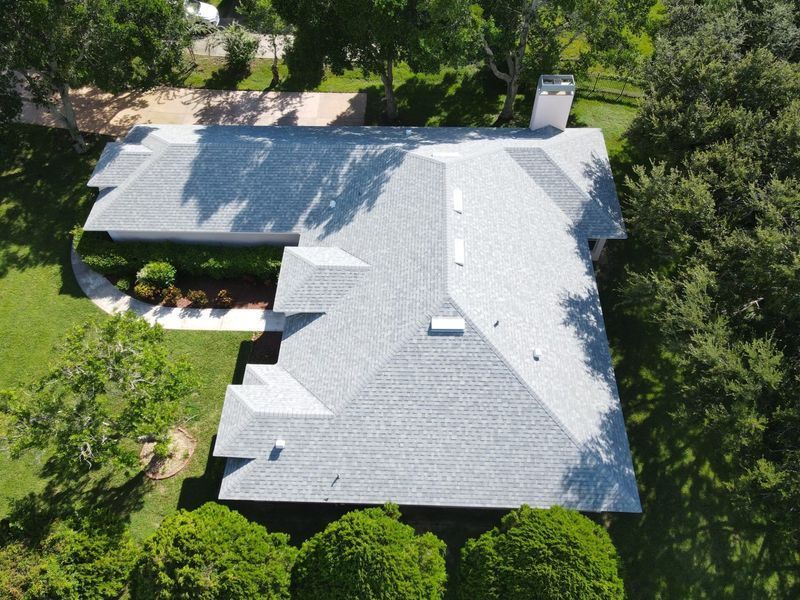 Aerial view of a light grey shingle roof on a house surrounded by green trees and grass.