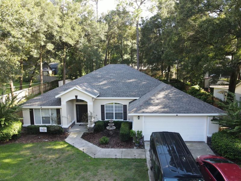 White house with gray roof, surrounded by trees and greenery, a black van parked in the driveway.