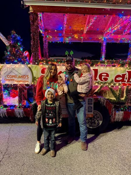 Family posing in front of a Christmas-decorated float at night. They are wearing festive sweaters.