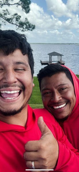 Two men smiling, giving a thumbs up, wearing red hoodies, by a lake.