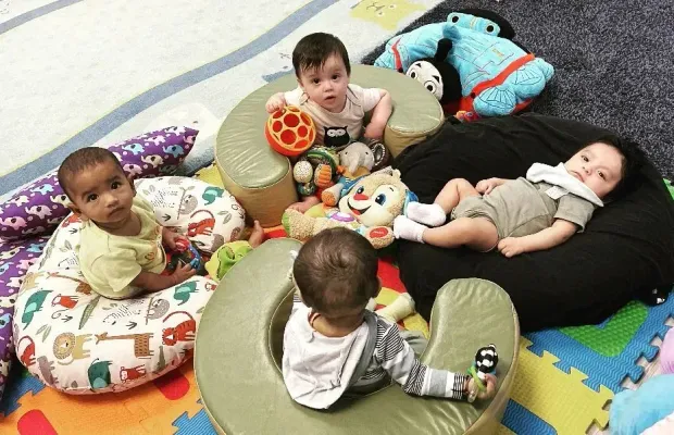 Babies sitting in various seats and playing with toys on a playroom floor.