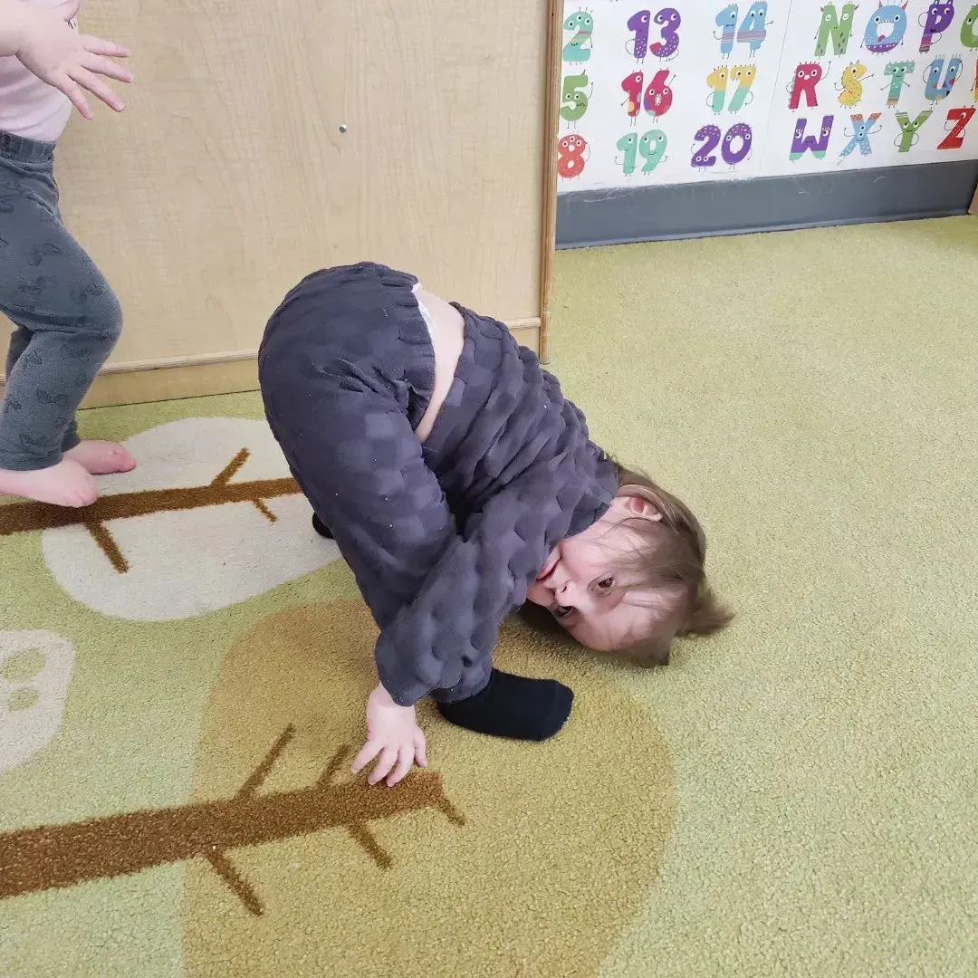 Child bent over, touching ground, wearing gray outfit. Another child's legs are visible at the left. Carpeted room with wall numbers.