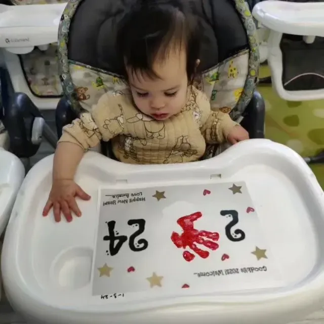 Baby in high chair with handprint on a paper, "24," on the tray.