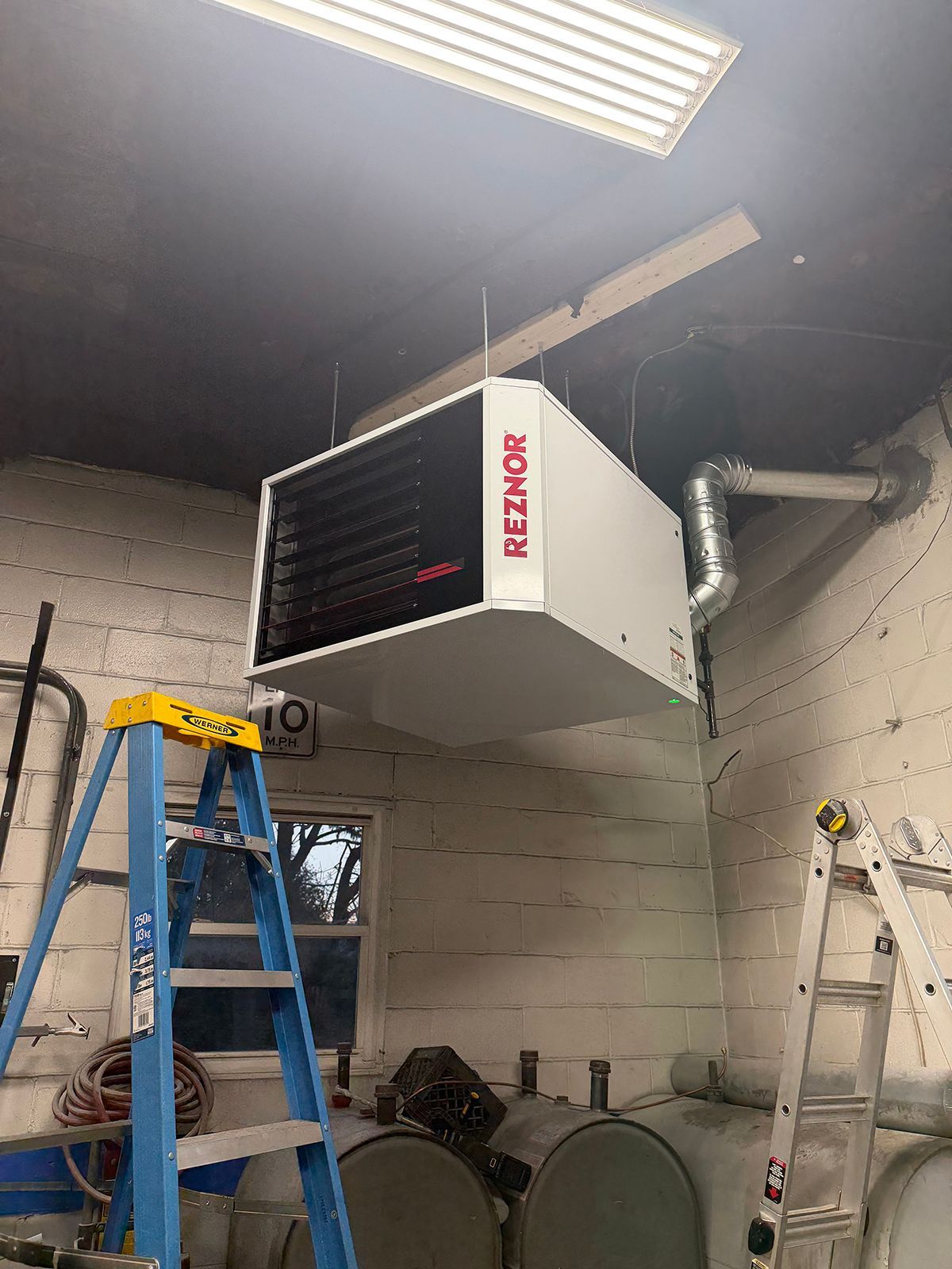 A Reznor unit heater suspended from the ceiling in a workshop with a blue ladder and a silver ladder underneath.